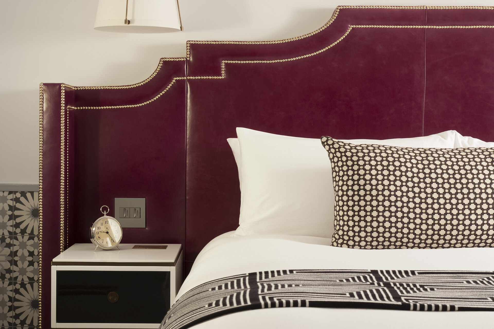 A close-up of a bed with a maroon headboard, white bedding, and patterned pillows.