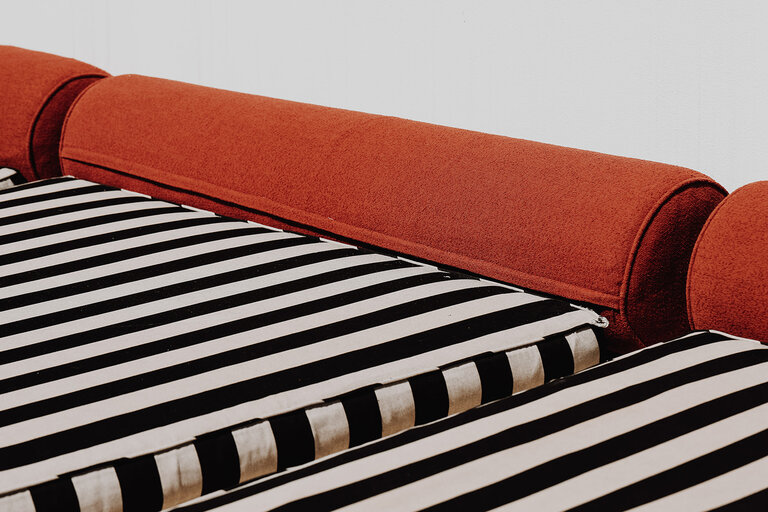 A close-up of a bed with black and white striped sheets and an orange bolster pillow.