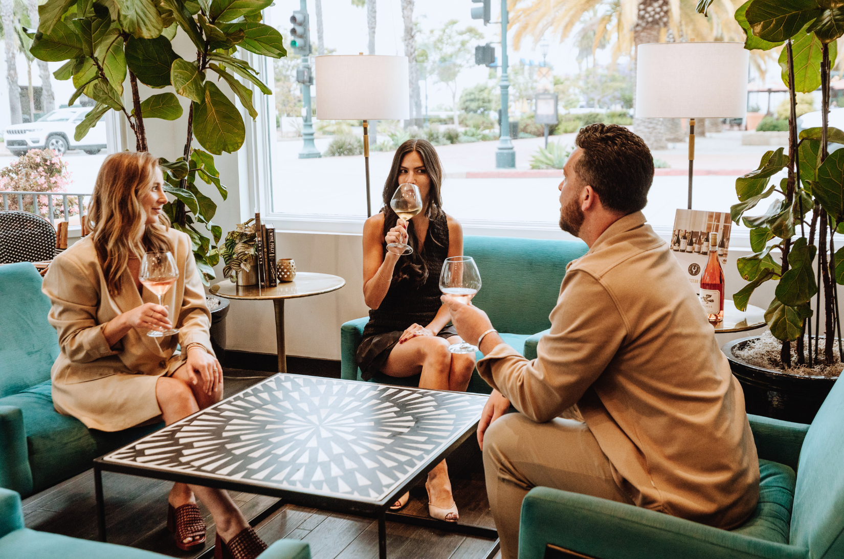 Three people sitting in a lobby lounge with teal sofas, a coffee table, and plants, enjoying wine.