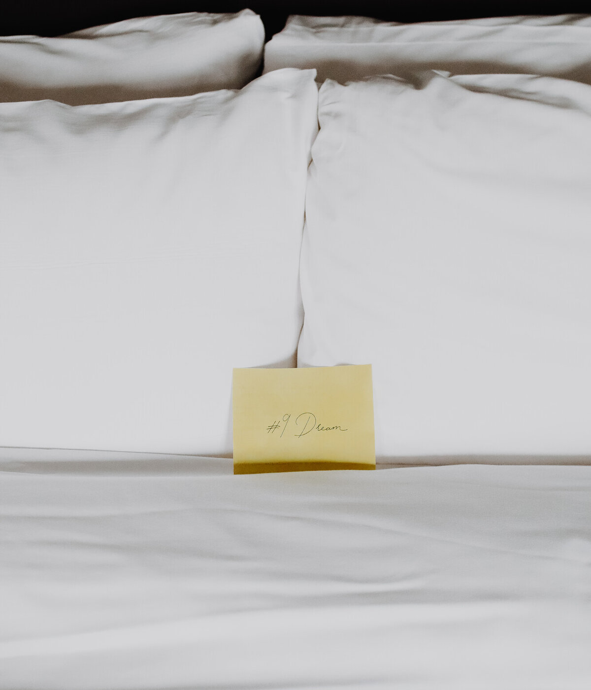 A close-up of a bed with white pillows and bedding, and a yellow note that reads '#9 Dream'