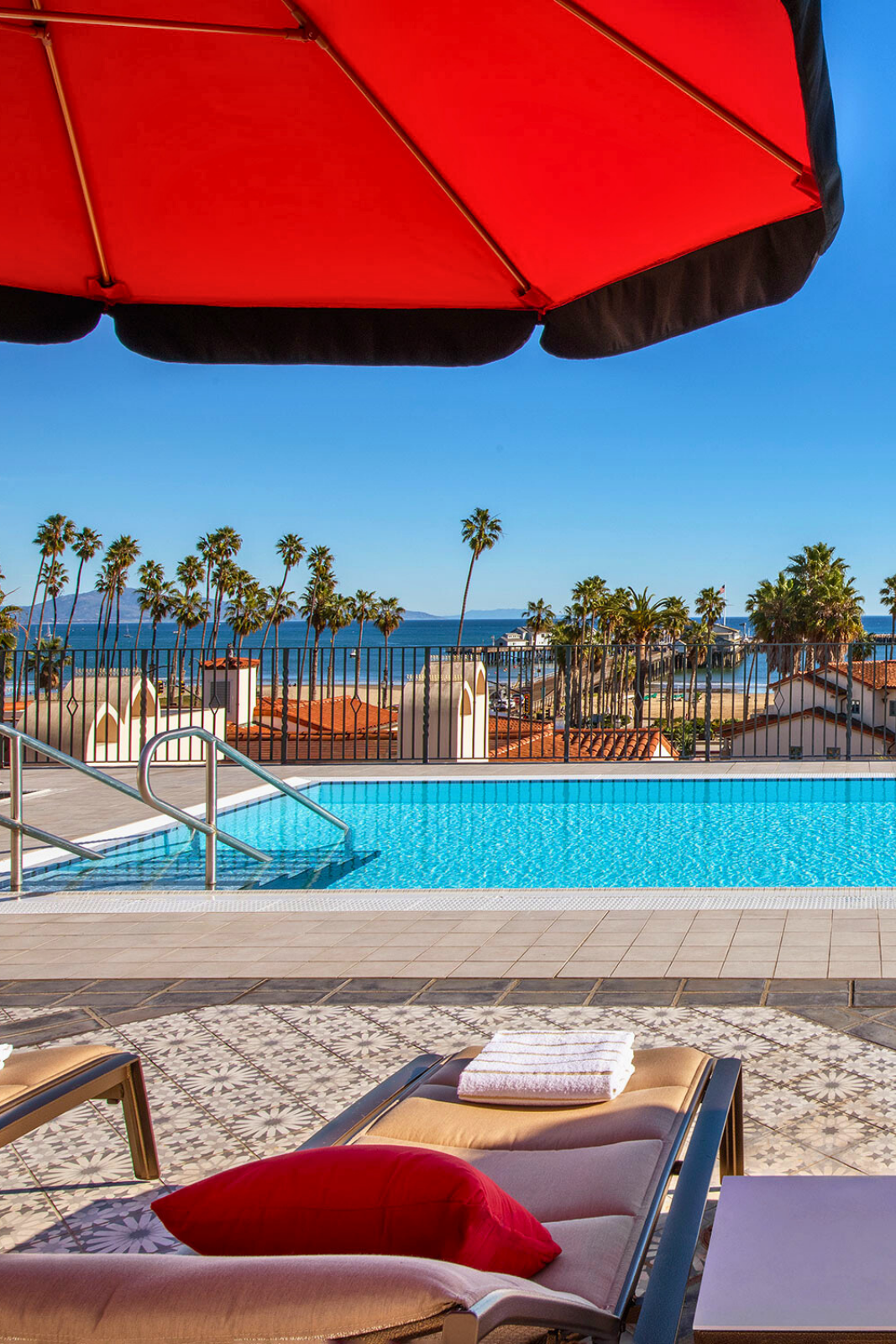 A pool area with a red umbrella, lounge chairs, and a view of the ocean and palm trees.